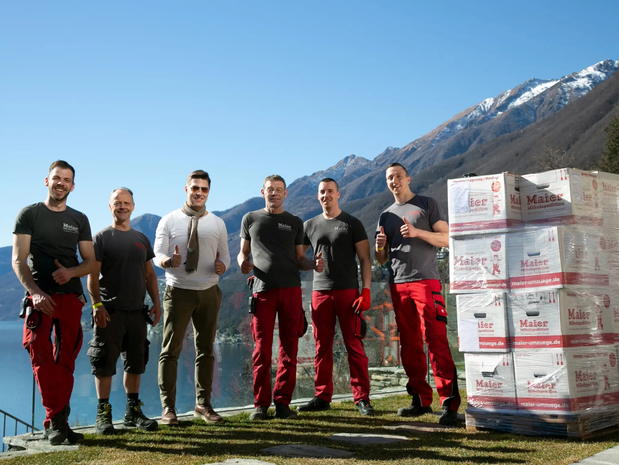 A group of smiling workers from Möbelspedition Maier stand proudly in work clothes with their customer in front of a large stock of moving boxes, with a picturesque mountain landscape in the background.