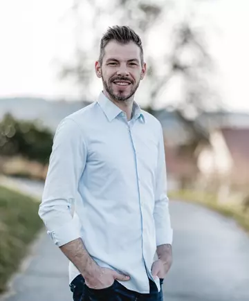 Dennis Maier the managing director of Möbelspedition Maier stands outside smiling, with a beard, in a light blue shirt and dark blue jeans with a blurred natural background.