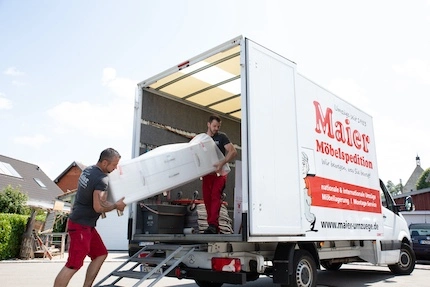 Two removal men unload a white piece of furniture from a removal van from Möbelspedition Maier e.K. on a sunny day.