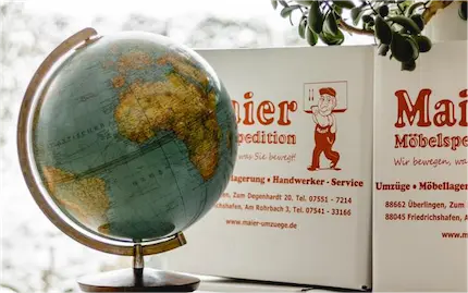 Ein Globus im Vintage-Stil neben einem offenen Karton mit Illustrationen und Texten in deutscher Sprache, in der Nähe eines Fensters, dessen Pflanzenblätter sanft ins Bild ragen. Der Karton enthält Gegenstände für einen Umzug.